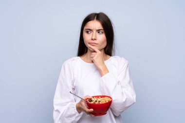 Young woman over isolated blue background holding a bowl of cereals and thinking