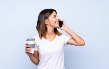 İzole edilmiş mavi arka planda, elinde kahve ve cep telefonu olan genç bir kadın.
