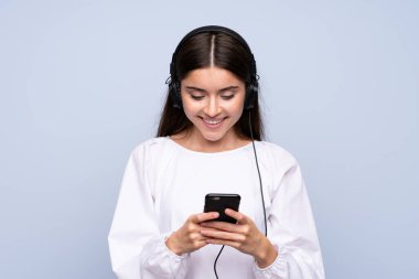 Mavi arka planda, kulaklıklı cep telefonunu kullanan genç bir kadın.