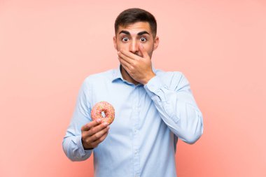 Sürpriz yüz ifadesi ile izole pembe arka plan üzerinde bir donut tutan genç adam
