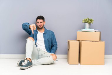 Yakışıklı genç adam kutuları arasında yeni bir evde hareket başparmak aşağı işareti gösteren