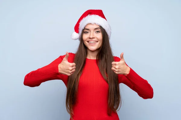 女の子とともにクリスマス帽子以上隔離された青背景与えます親指アップジェスチャー — ストック写真