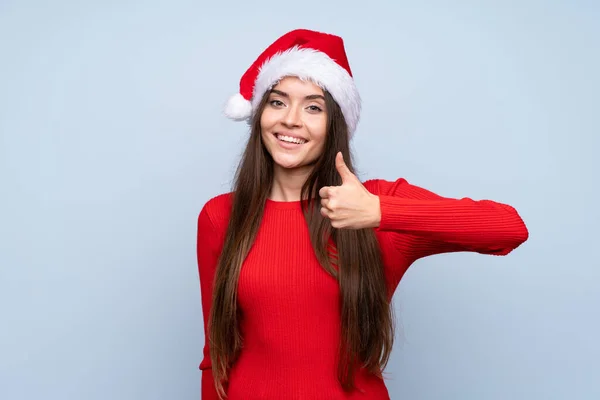 戴著圣诞帽 戴着蓝色背景的女孩举起了大拇指 — 图库照片