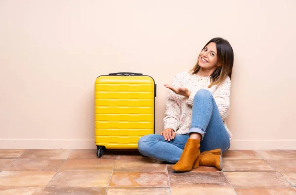 Traveler woman with suitcase sitting on the floor extending hands to the side for inviting to come