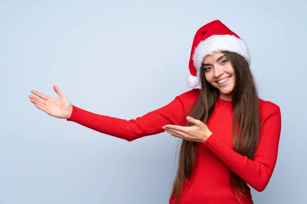 带着圣诞帽 披着蓝色孤立背景的女孩向旁边伸出手来 以感谢她的邀请 — 图库照片