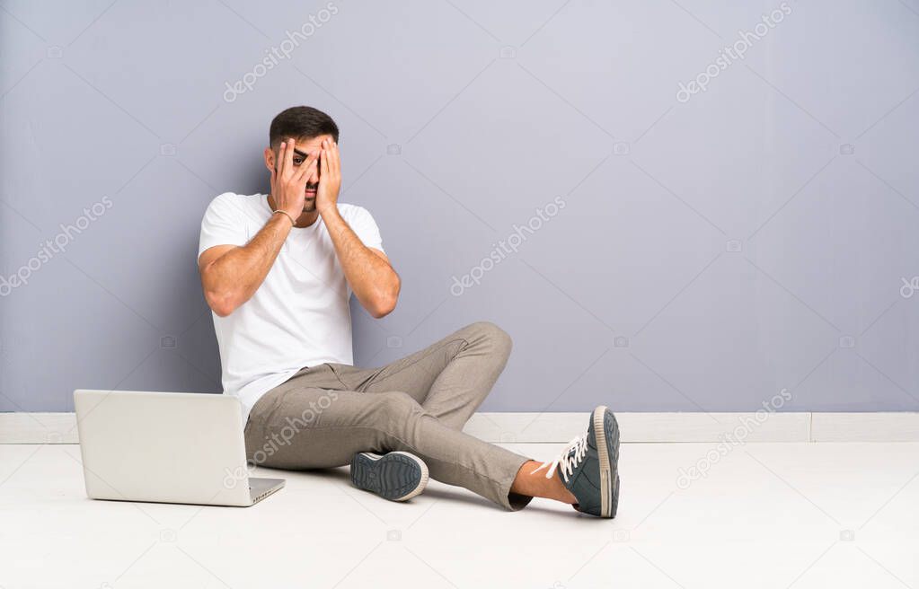 Young man with his laptop sitting one the floor covering eyes and looking through fingers