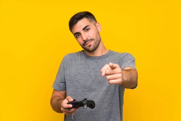 年轻英俊的男子在孤立的黄色背景下玩电子游戏控制器 带着自信的表情指着你 — 图库照片