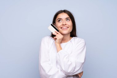 Soyutlanmış mavi arka planı olan genç bir kadın kredi kartı tutuyor.