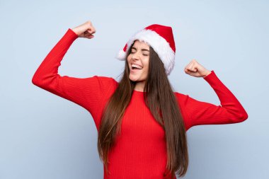 İzole edilmiş mavi arka planda Noel şapkalı kız zaferi kutluyor.