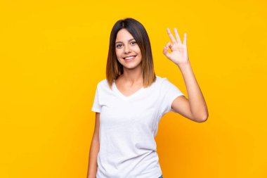 İzole edilmiş sarı arka plan üzerinde genç bir kadın parmaklarıyla tamam işareti gösteriyor.
