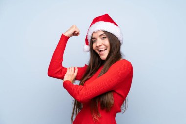 İzole edilmiş mavi arka planda, Noel şapkalı kız güçlü bir jest yapıyor.