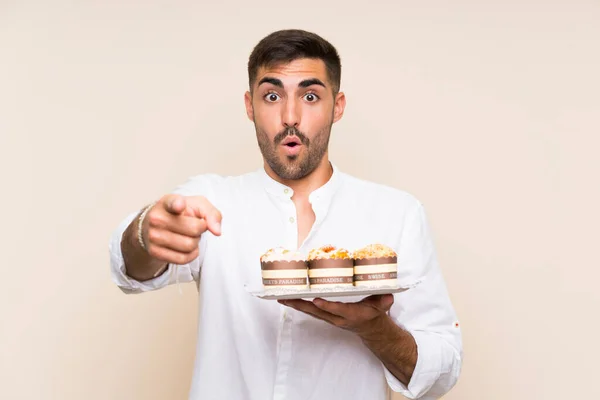 Handsome man holding muffin cake over isolated background surprised and pointing front