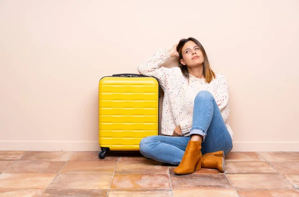 Traveler woman with suitcase sitting on the floor having doubts and with confuse face expression