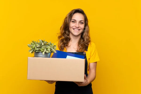 Young blonde girl making a move while picking up a box full of things smiling a lot