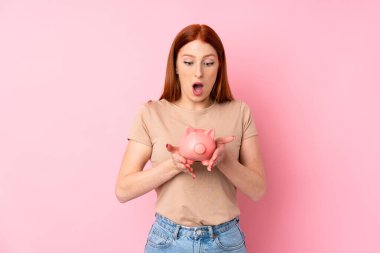 Soyutlanmış pembe arka planı olan genç kızıl saçlı kadın elinde büyük bir kumbara tutuyor.