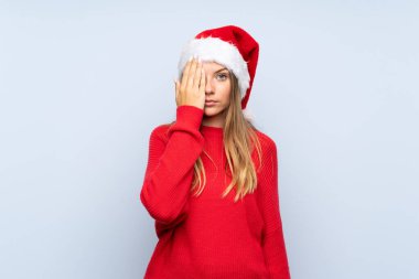 İzole edilmiş mavi arka planda Noel şapkası takan bir kız, bir gözü elle kapatıyor.