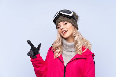 Snowboard gözlüklü kayakçı kız izole edilmiş mavi arka planda parmağını yan tarafa gösteriyor.