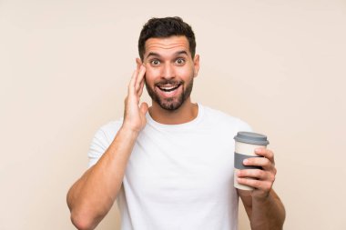 Sakallı genç adam, mavi arka plan üzerinde kahve tutuyor. Şaşırmış ve şok olmuş bir yüz ifadesi var.