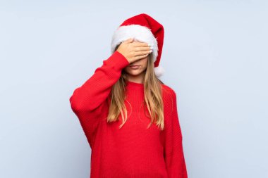 İzole edilmiş mavi arka plan üzerinde Noel şapkası olan kız gözlerini elleriyle kapatıyor.