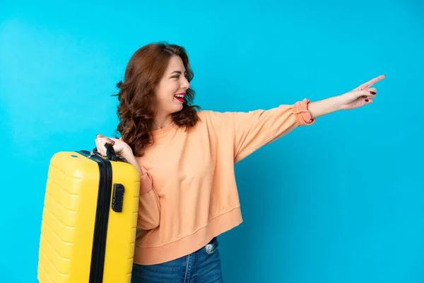 Traveler woman with suitcase over isolated blue background pointing finger to the side