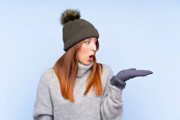Young redhead Russian woman with winter hat over isolated blue background holding copyspace imaginary on the palm