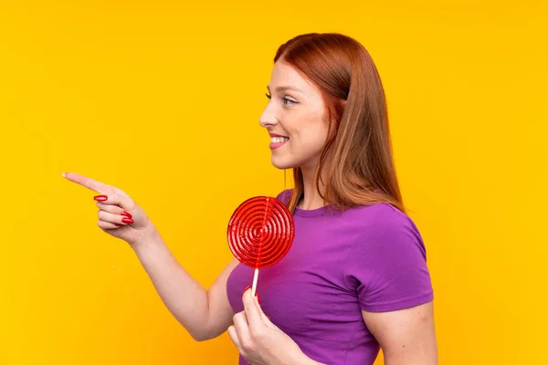 Young Redhead Woman Holding Lollipop Yellow Background Pointing Side Present — Stock Photo, Image