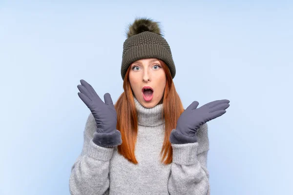 Young redhead Russian woman with winter hat over isolated blue background with surprise facial expression