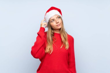 İzole edilmiş mavi arka plan üzerinde noel şapkası olan kızın şüpheleri ve kafa karıştırıcı bir yüz ifadesi var.