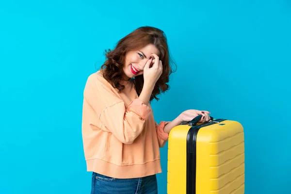 Traveler woman with suitcase over isolated blue background laughing