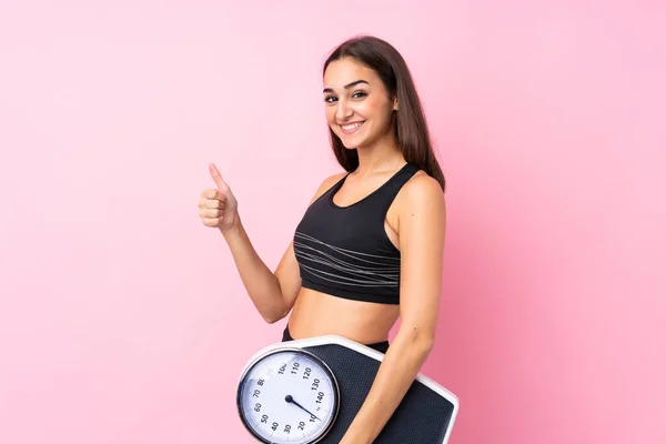 Pretty young girl with weighing machine over isolated pink background holding weighing machine with thumb up