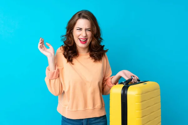 Traveler woman with suitcase over isolated blue background laughing