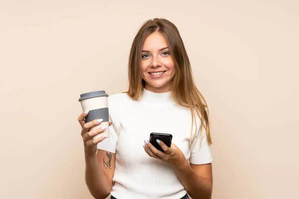 一个金发碧眼的年轻女人 背景偏僻 手里拿着咖啡和手机 — 图库照片