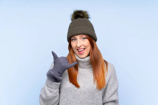 若いですRedheadロシア人女性とともに冬の帽子以上隔離された青背景作る電話ジェスチャー — ストック写真