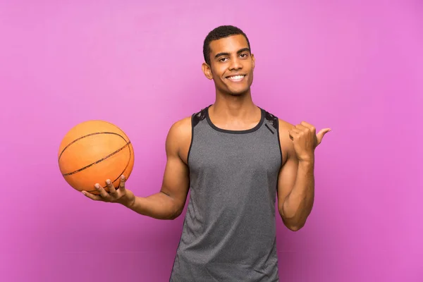 Молодой Спортсмен Мячом Баскетбола Над Изолированной Фиолетовой Стеной Указывая Сторону — стоковое фото