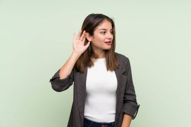 Yeşil arka planda ceketi olan genç esmer kız elini kulağına koyarak bir şeyler dinliyor.