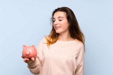 Genç esmer kız izole edilmiş mavi arka planda büyük bir kumbara tutuyor.