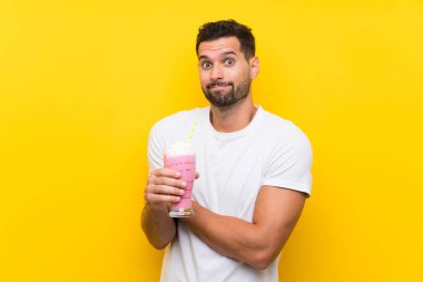 İzole edilmiş sarı arka planda çilekli milkshake 'i olan genç adam omuzlarını kaldırırken şüpheyle hareket ediyor.