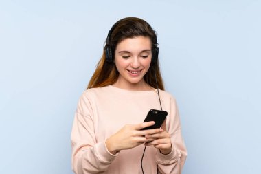 Genç esmer kız mavi arka planda kulaklıklı cep telefonunu kullanıyor.