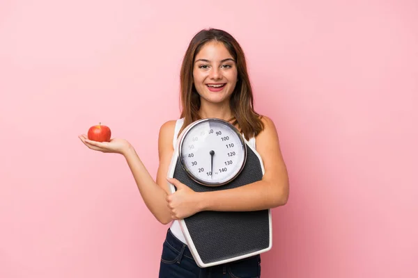 Young pretty girl with weighing machine and with an apple over isolated pink background