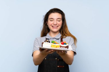 Genç esmer kız izole edilmiş mavi arka planda mini pastalar tutuyor.