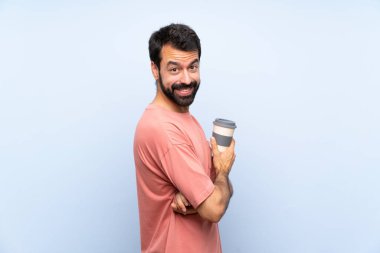 Sakallı genç adam izole edilmiş mavi arka planda kahve tutuyor. Kolları çapraz ve ileriye bakıyor.