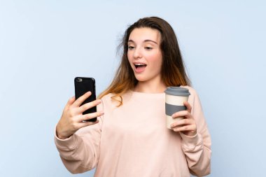 Genç esmer kız izole edilmiş mavi arka planda elinde kahve ve bir cep telefonu var.