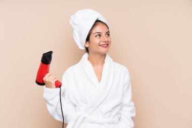 Üzerinde saç kurutma makinesi olan bornozlu genç bir kadın izole bir arka planda gülümserken yukarı bakıyor.