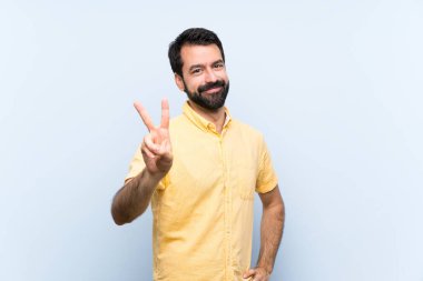 Mavi arka planda sakallı genç adam gülümsüyor ve zafer işareti gösteriyor.