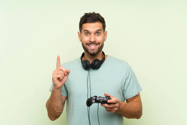 素晴らしいアイデアを指し示す孤立した緑の壁の上にビデオゲームコントローラで遊ぶ若いハンサムな男 — ストック写真