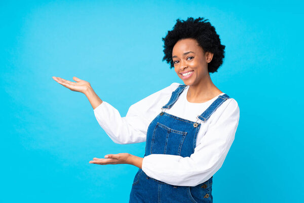 African american woman with overalls over isolated blue background extending hands to the side for inviting to come
