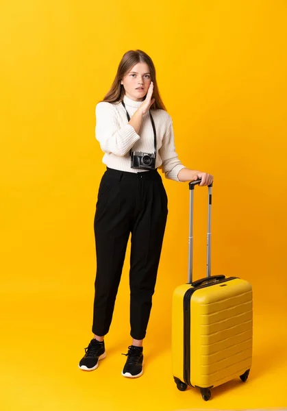 Full body of traveler teenager girl with suitcase over isolated yellow background whispering something