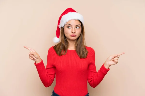 年轻姑娘头戴圣诞礼帽 背负孤立的背景 指向有疑问的侧面 — 图库照片