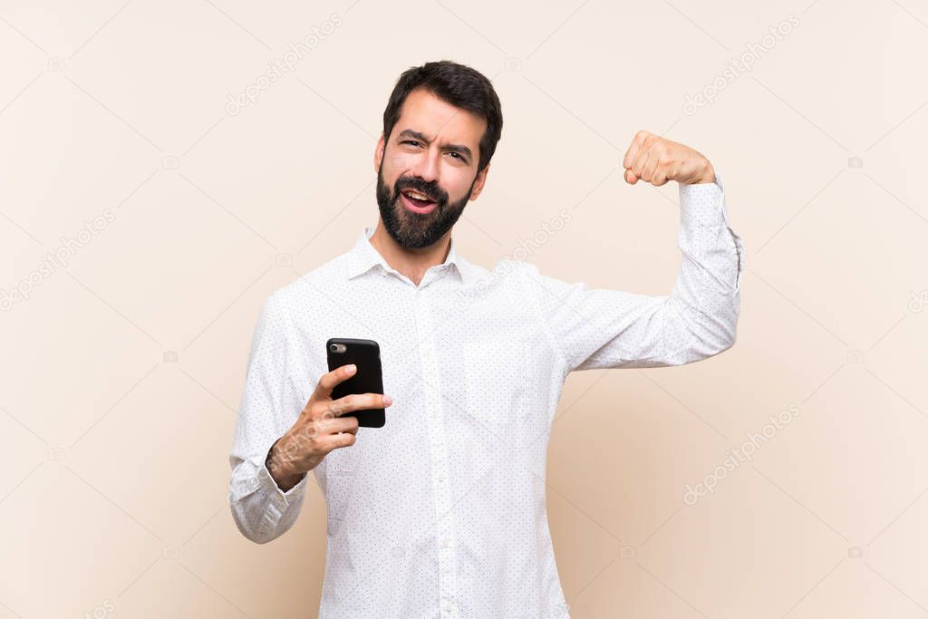 Young man with beard holding a mobile doing strong gesture