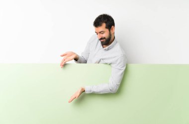 Sakallı genç yakışıklı adam, büyük yeşil boş bir pankart tutuyor. Gelmesi için ellerini yana uzatıyor.